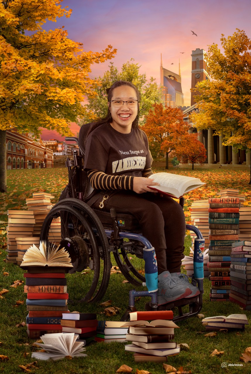 a young Asian woman is seated in her wheelchair on a leafy lawn meant to represent Vanderbilt’s campus, wearing a “Next Steps at Vanderbilt” T-shirt. The Batman building, a key part of the Nashville skyline, is behind her in the distance. She has long black hair, glasses and a big smile and has a book open on her lap. Stacks and stacks of closed and open books lay around her on the grass. 