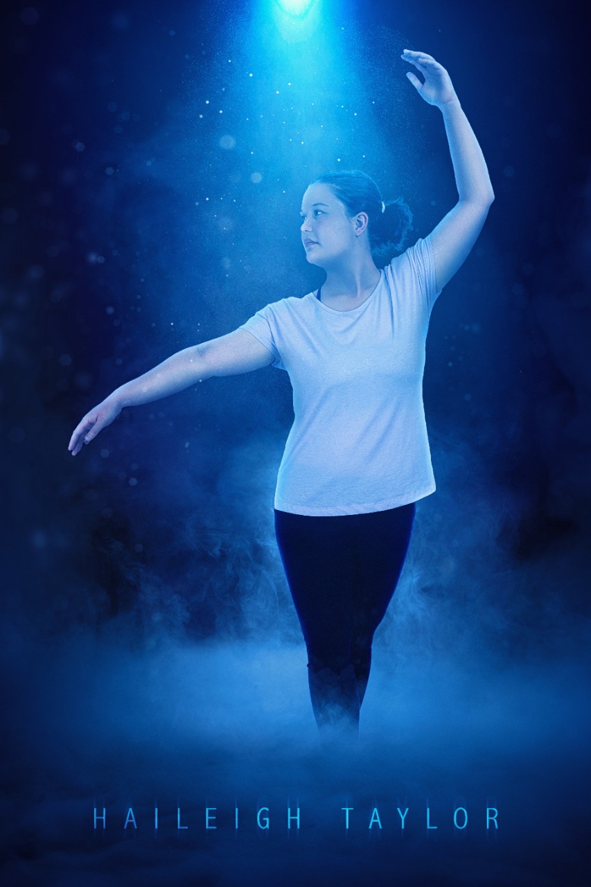 a young white woman in a white t-shirt, black leggings and her brown hair pulled back in a ponytail poses in a dance move with one arm arched gracefully over her head and the other arm out to the side of her body. She’s shown in a blue misty spotlight