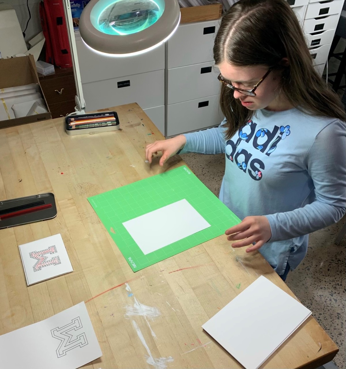 Marie is a teen with Down syndrome, long brown hair, glasses and a blue shirt. She stands and looks down at a worktable with two pieces of paper on it.