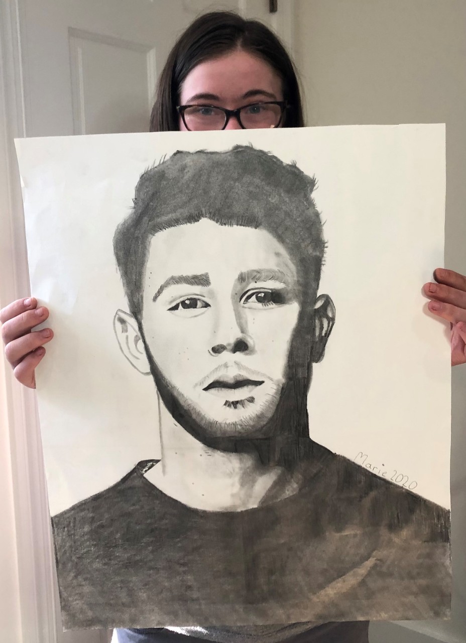 Marie holds up a large canvas with an incredibly impressive and detailed, lifelike sketch of the pop singer Nick Jonas in black and white, shown from the chest up. Marie’s face with brown hair and glasses just peeks over the top of the large piece of art. Marie has Down syndrome.