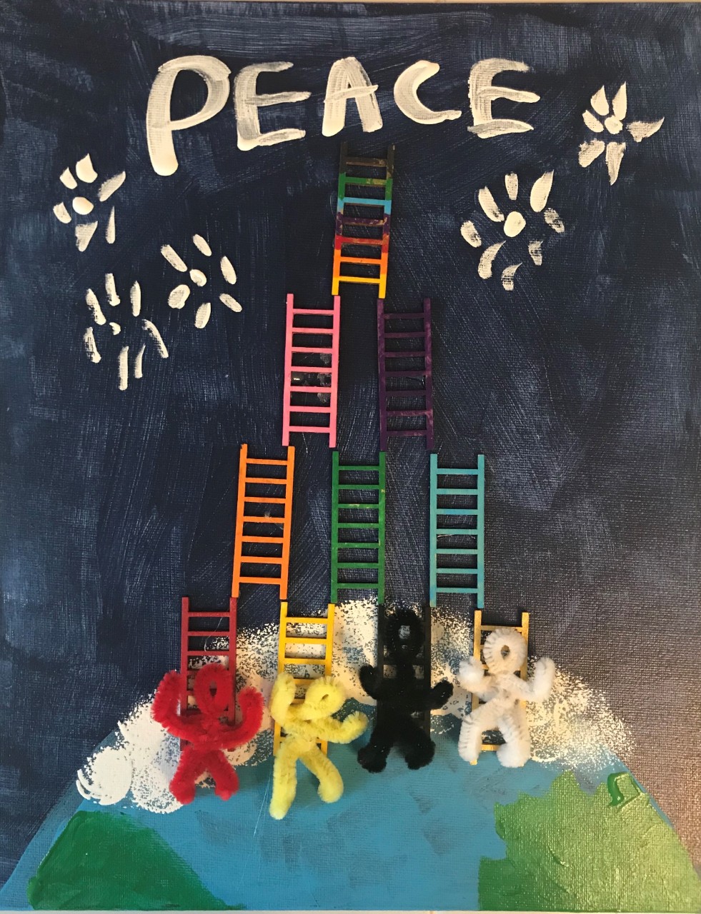 A mixed-media piece. The canvas is painted blue and the Earth is painted at the bottom of the piece. There are figures of people shown with different colored pipe cleaners glued onto the Earth, climbing an array of tiny colorful wooden ladders also glued to the canvas.  They are climbing up to the top of the canvas where the word “Peace” is written in white paint.