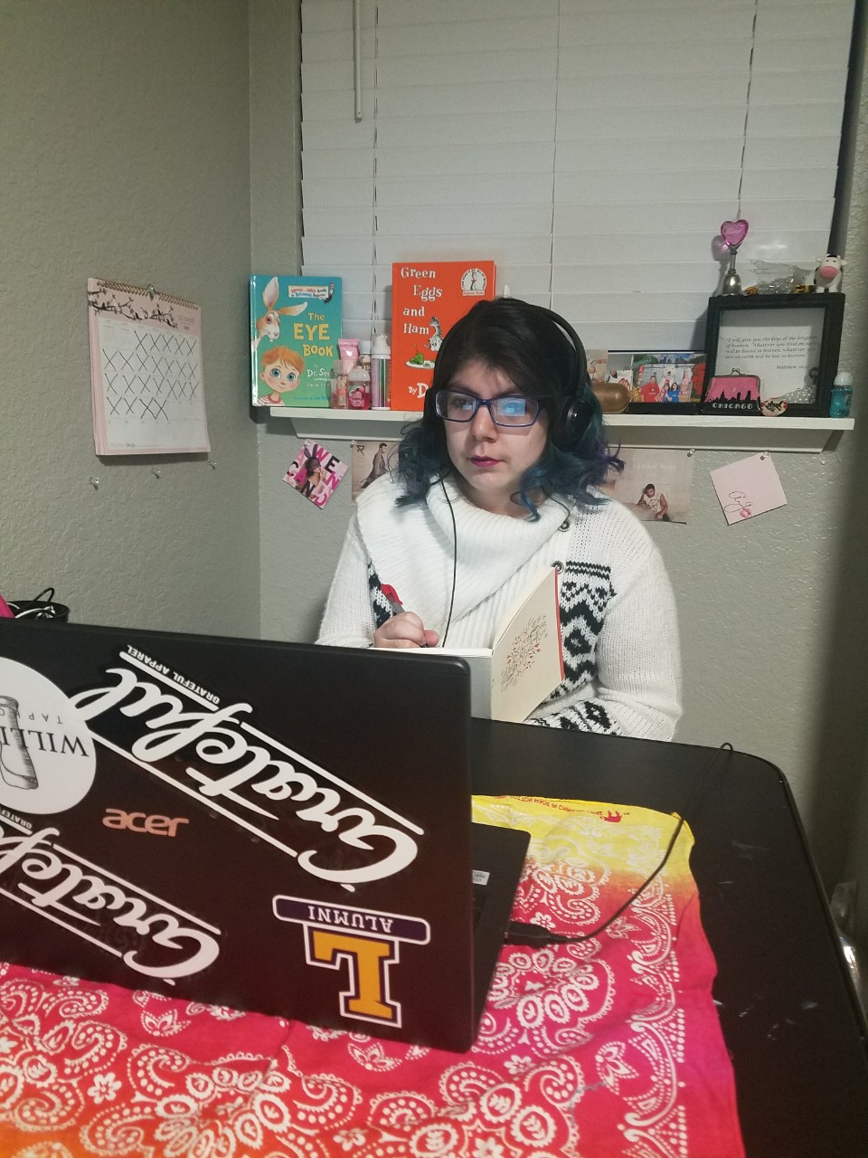 a young Latina woman sitting at a table in her home, looking at her laptop that is covered in stickers. She is wearing a big cozy white sweater and listening to headphones plugged into the computer.