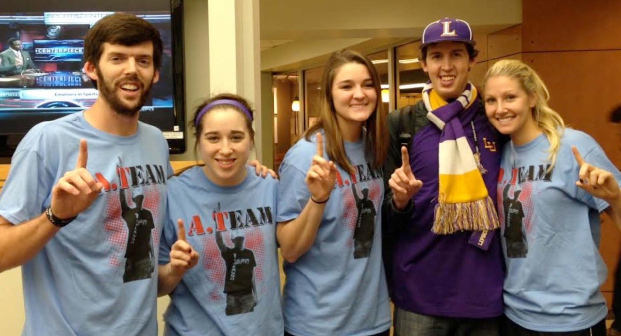 Lipscomb IDEAL program and shows 5 white college students, two boys and three girls with and without intellectual disabilities. 4 are in matching T-shirts that say “A-Team” and the other young man is wearing Lipscomb sports colors of purple and gold. They are all holding up 1 finger in a kind of salute and smiling