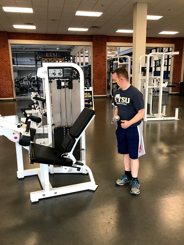 Garrison Buchanan cleans the fitness equipment. A Young white man with Down syndrome dressed in athletic gear wipes down fitness equipment in a gym.
