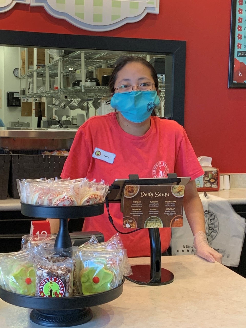 Union student Faith wearing a bright pink shirt and standing behind the front counter at the restaurant where she works, in front of the cash register. She’s wearing a mask and a name tag.