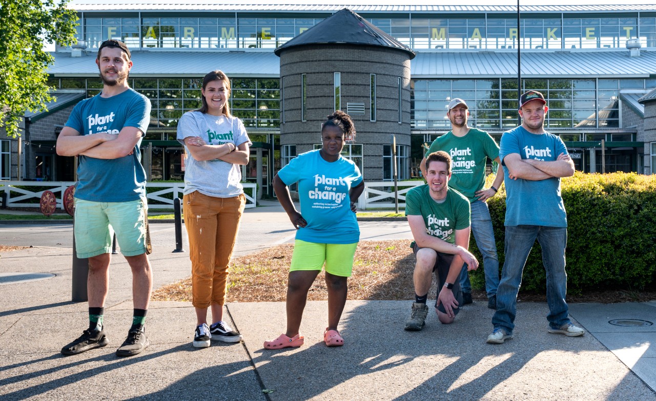 group photo of people with and without disabilities – two young women, and four young men. They are posed standing and kneeling outside of the Nashville Farmer’s Market, where Plant for a Change hosts a plant selling stand for their business. They are all wearing matching T-shirts that say “Plant for a Change.”