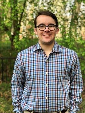 Wilson, a young white man with glasses, short brown hair and a button up blue plaid shirt stands outdoors and smiling for the posed photo