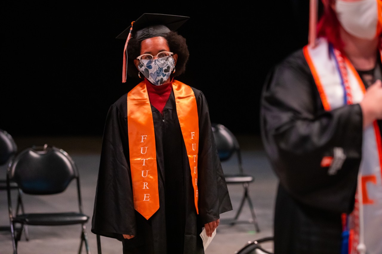 a young Black woman, Ciara Armstrong, a UT FUTURE graduate in her graduation gown with a bright orange sash around her neck that says “FUTURE”. She is also wearing a graduation cap and tassel, and mask.