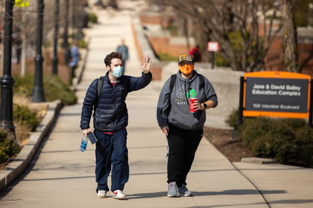 two young men walking outside on campus with masks on and backpacks. One is waving to someone off camera.