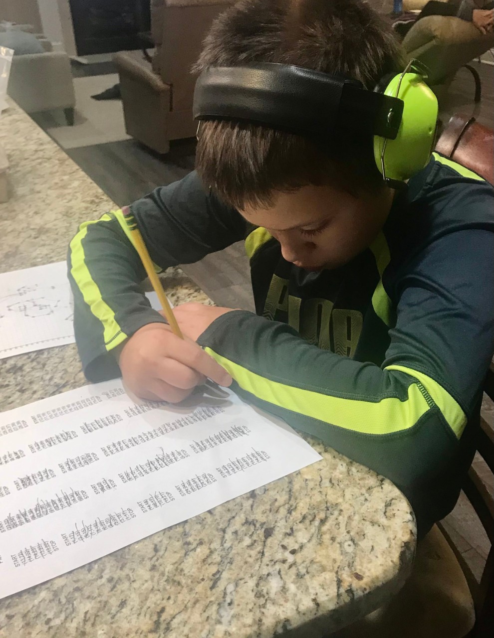 a young boy with brown hair wearing big noise-canceling earphones sits at a table and looks down at his homework intently writing with a pencil. 
