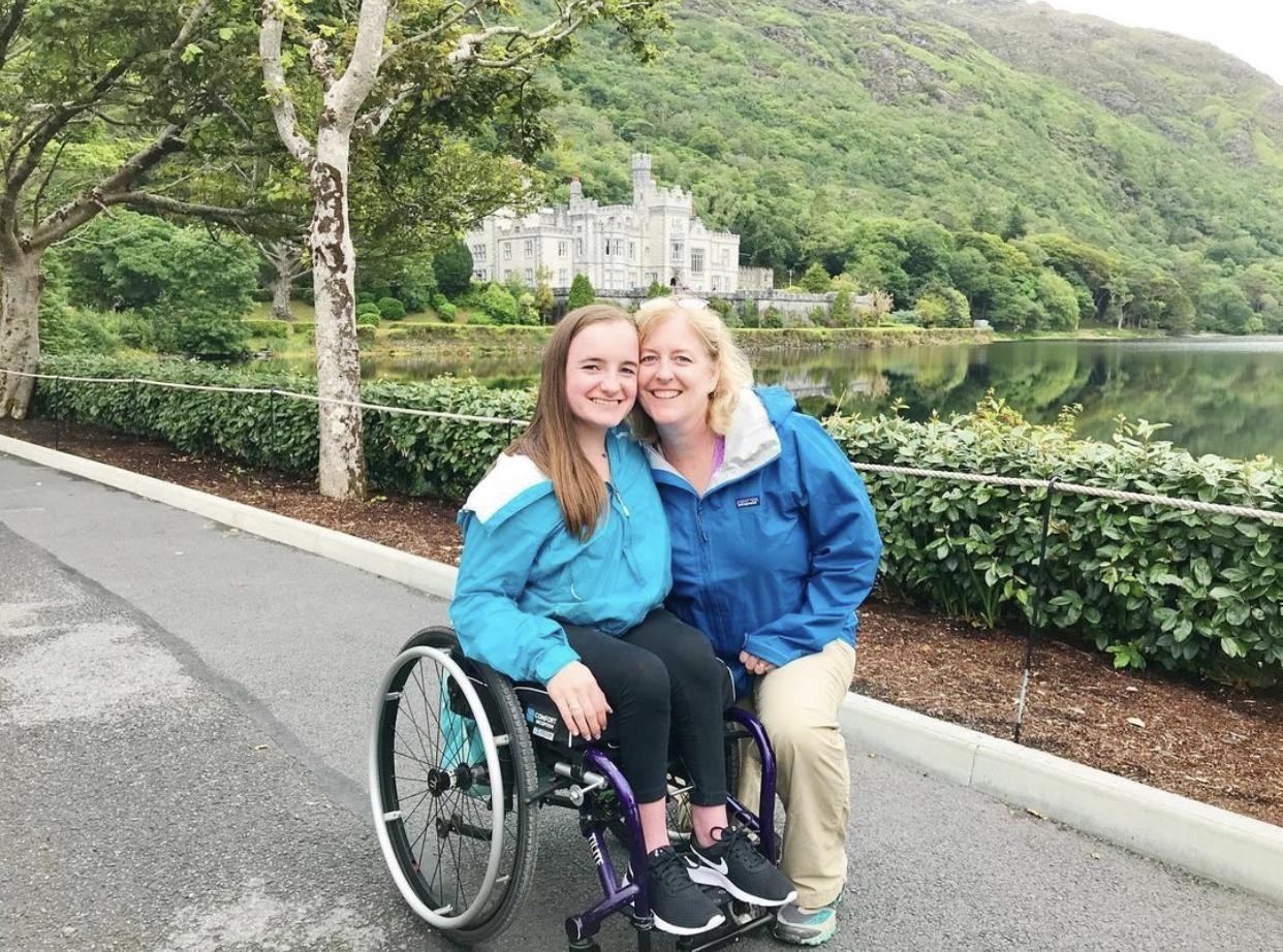 Rosie and her mom, Carol, pose in front of Kylemore Abbey on the west coast of Ireland. Rosie’s mom is crouching down to pose next to her daughter. There is a majestic building, the abbey, behind them surrounded by trees. Both of them are wearing blue rain jackets and smiling.