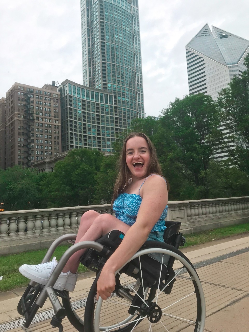 Rosie on a visit to Chicago, Illinois. She is smiling really big and tilting back in her wheelchair a bit. There are big buildings behind her. She is in a bright blue patterned tank top and shorts.