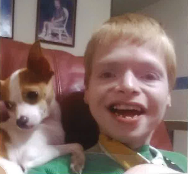 Jeffery Lambdin is a close-up photo of a white man with red hair with a visible disability and big smile; he has a cute little brown and white dog sitting on his shoulder as he leans back against a couch