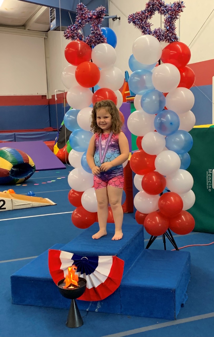 a young girl with curly brown hair wearing a leotard stands in a gym on a block wearing a medal like she has won a prize for gymnastics; she has a big smile on her face and there are red, white and blue balloons behind her