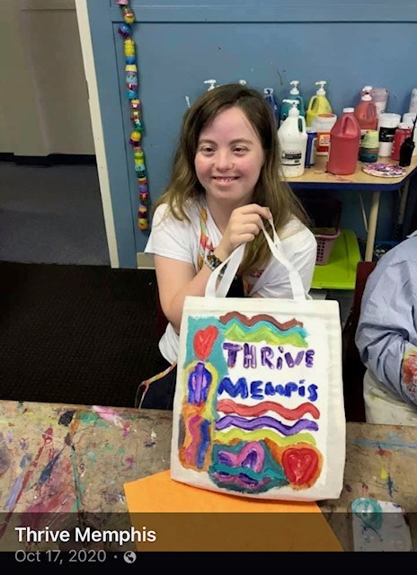 A young woman with Down syndrome with a big smile holds up a tote bag she just painted with all sorts of colorful designs, with art supplies behind her.