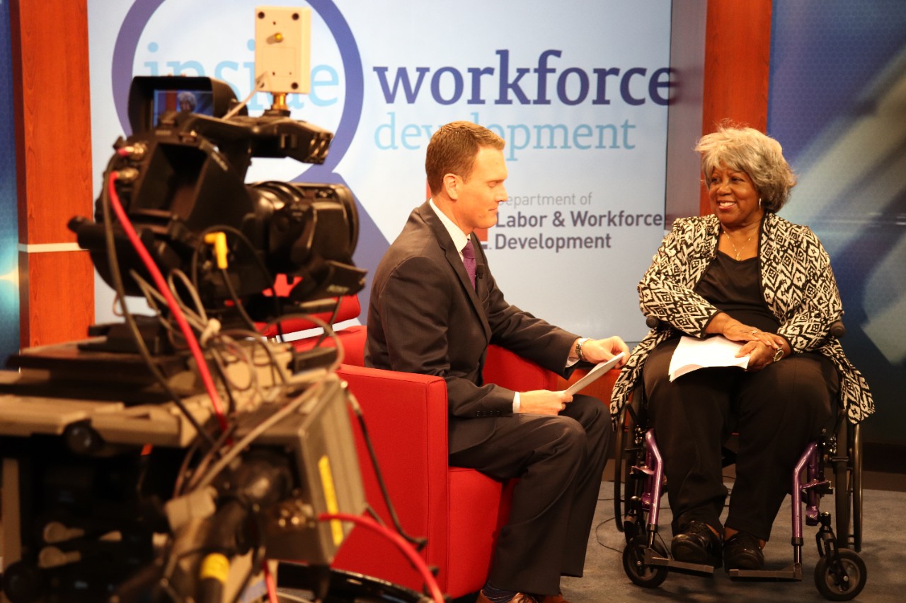 Two people are seated on a TV set. A white man in a suit sits on a red chair. A Black woman is seated in a wheelchair. The screen in the background reads, “Workforce Development.” A large camera rig is in the foreground in the left-hand corner.