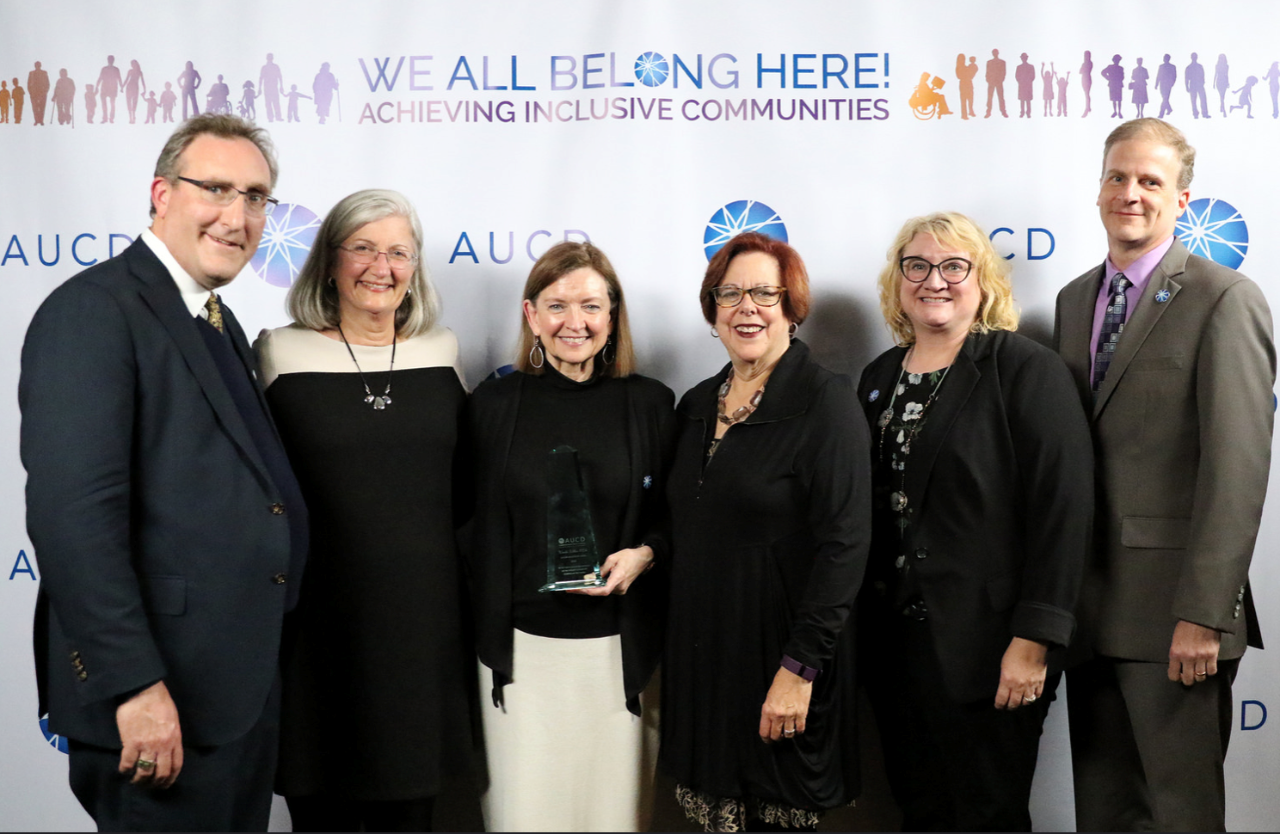 Six people in dark dress clothes pose in front of an event background. The banner says at the top, “We all belong here! Achieving Inclusive Communities.” Wanda is in the center of the group, with a white man and a white woman with gray hair on the left, and two white women and a white man on her left. Wanda is holding a clear award