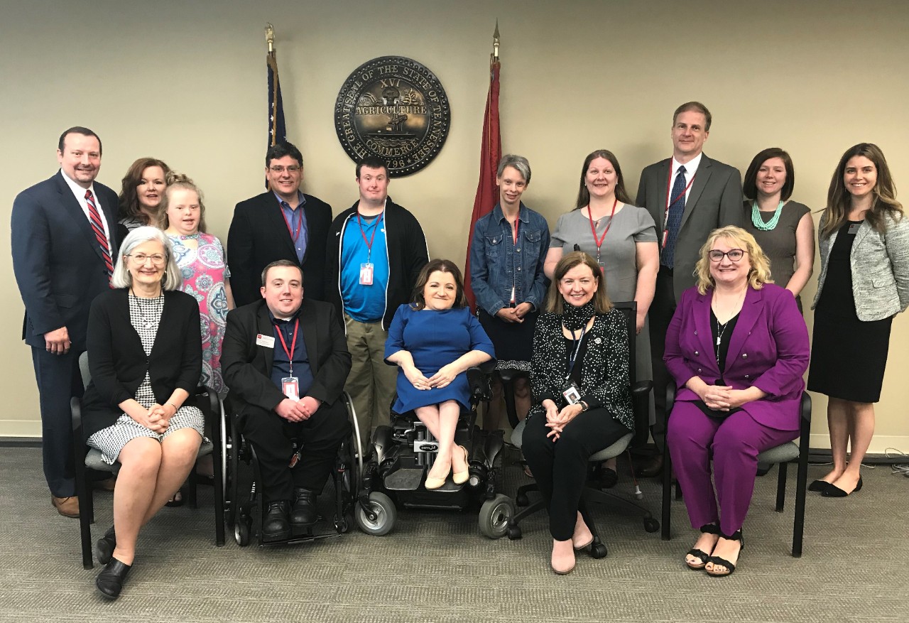 A group of 15 people pose in front of the TN state seal and U.S. and Tennessee flags. The people are all different ages, both men and women, dressed in business wear. The group includes people with different visible disabilities, including Down syndrome, Williams syndrome, and two individuals in the front row who use wheelchairs. 