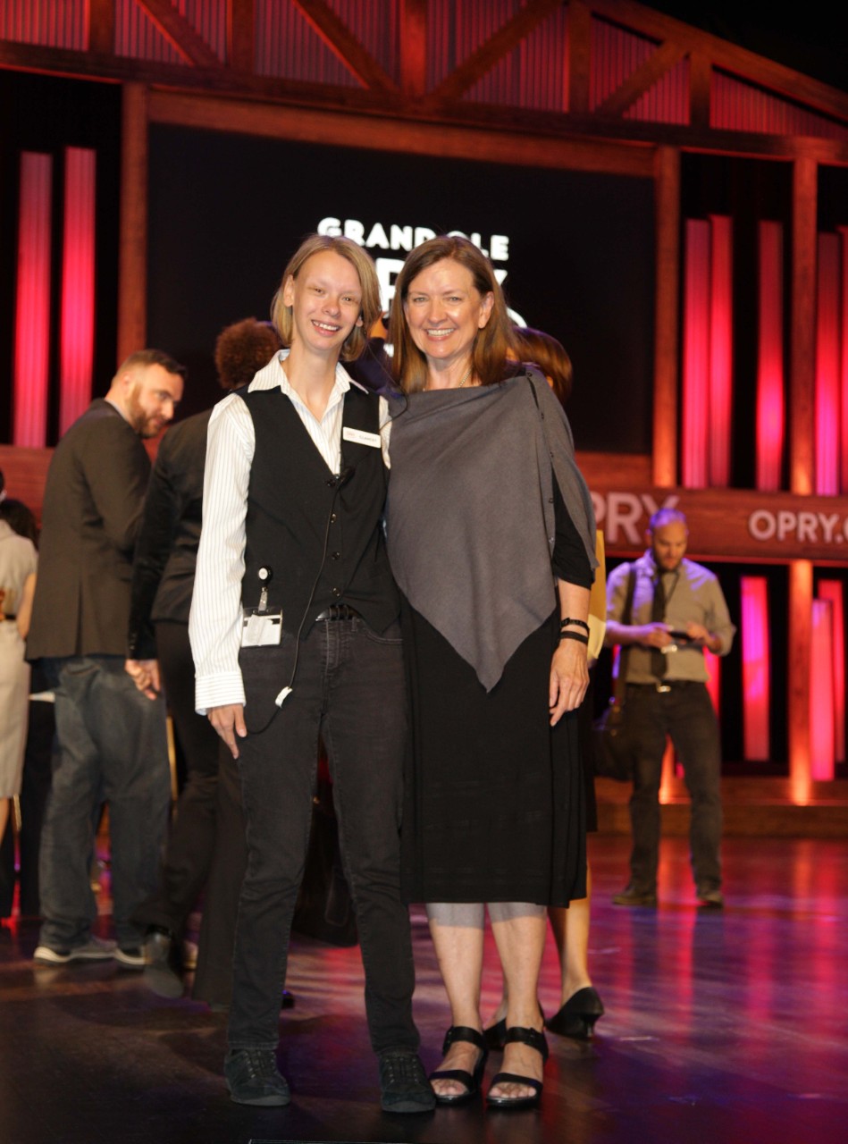 Wanda, wearing a black dress and grey shawl, has her arm around Clancey, a young woman with Williams syndrome in an Opry tour guide uniform, and they are both smiling on the Grand Ole Opry stage, with other people in the background