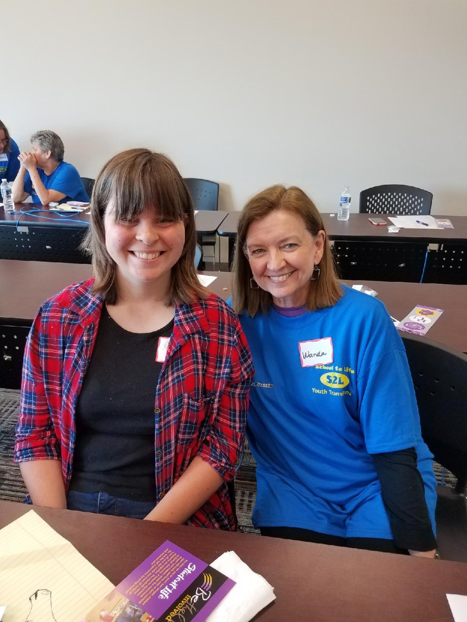 Wanda and a young woman with a disability sit beside each other in a classroom; Wanda is wearing a bright blue shirt representing the youth leadership program she is visiting in Paris, TN