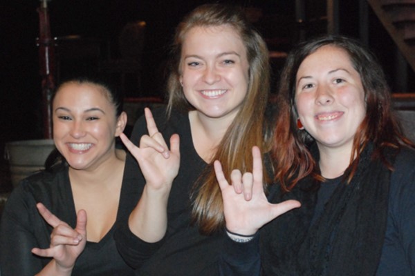 3 young women wearing all black pose together and smile for a photo while signing in sign language “I love you.”