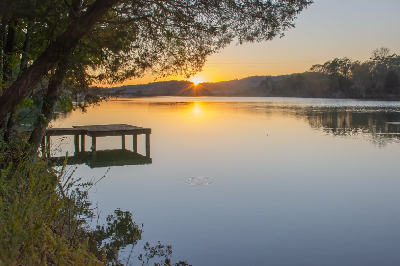 A photo of the sun setting on a body of water with a wooden dock emerging from the bank on one side and hills and forests in the background on the other side of the water.