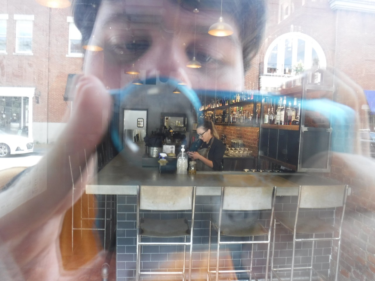 This photo shows the reflection of a white young man taking a photo with a camera in the window of a coffee shop. In the center of the photo there is a barista at the coffee counter preparing a drink but around the image of what is inside the shop is the photographer’s reflection while he is concentrating on taking the digital photo.