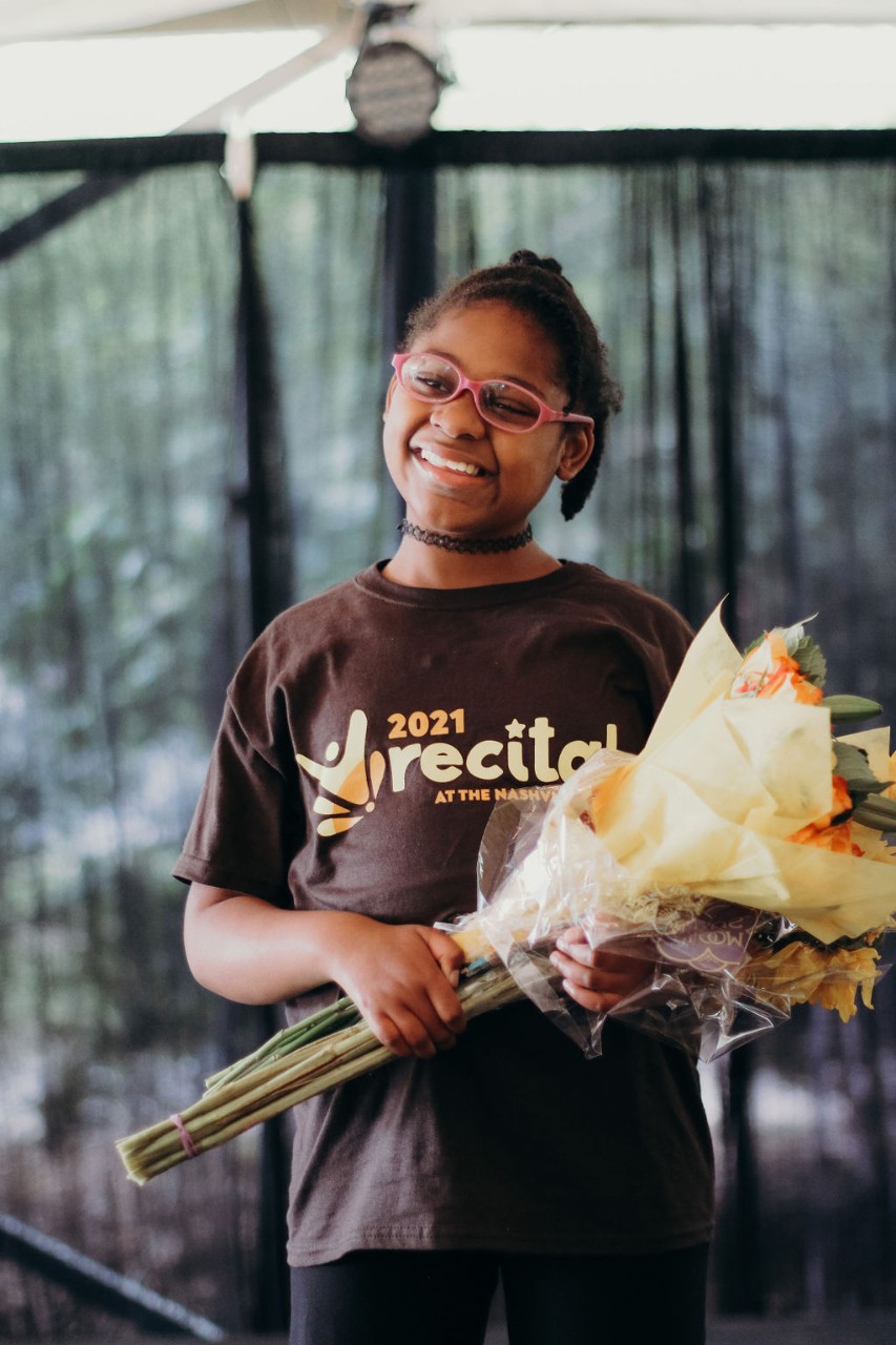 A young Black girl wearing pink glasses and a “2021 recital” T-shirt poses smiling while holding a bouquet of flowers