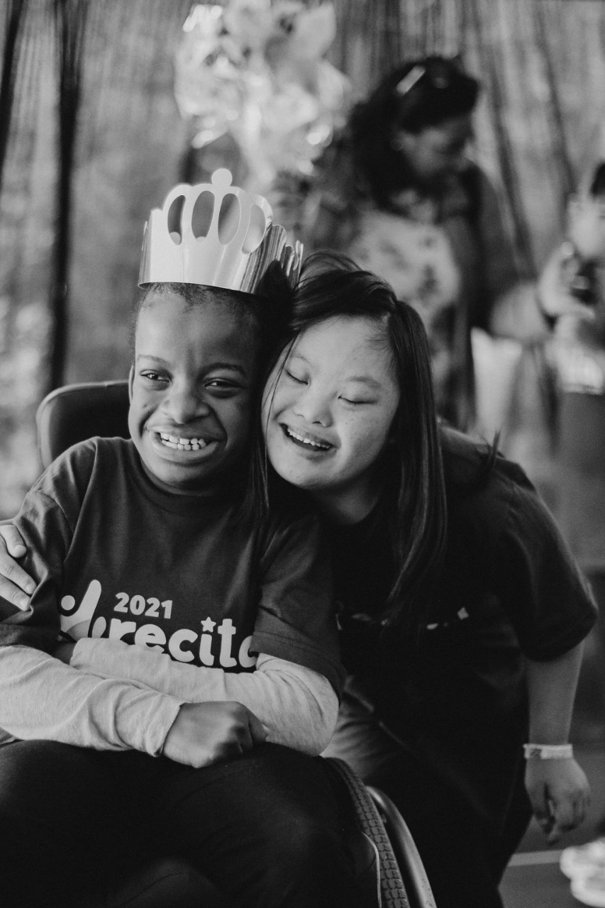 A young Black girl in a wheelchair wearing a paper crowd and “2021 recital” shirt is hugged from the side by her sister, a young Asian girl with a huge smile.