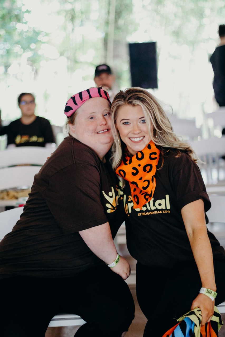 Two young white women lean together for a photo in matching black T-shirts for the recital. One of the women has Down syndrome, both are also wearing colorful bandanas.