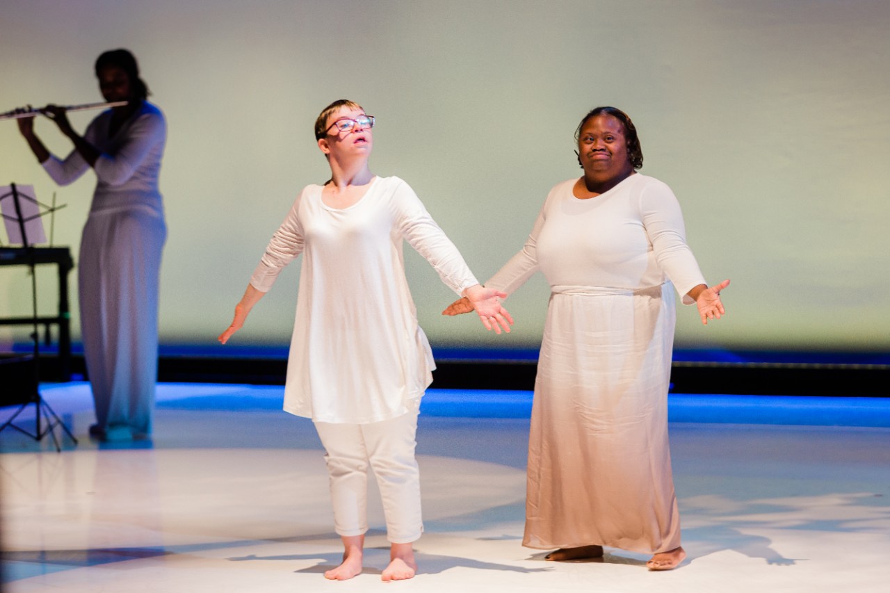 Friend’s Life participants and dancers Kaylea Dunkin and Keonna Reed are shown standing side by side centerstage with their arms spread out. They are wearing loose and flowing white clothing in front of a white backdrop. Behind them in the shadows you can see a flute player playing the music they are dancing to. Kaylea is a white young woman with Down syndrome with short blonde hair and glasses. Keonna is a Black young woman with Down syndrome with short black hair and a big smile