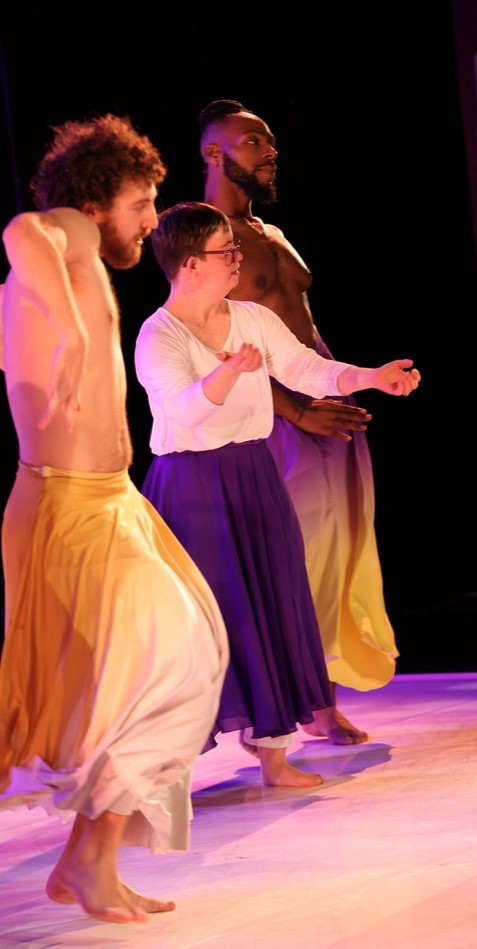 3 dancers, two men and a woman, are shown from the side standing in a row in the midst of performing. The woman is a young woman with Down syndrome with short cropped blonde hair, a loose white blouse, and a long flowing purple skirt. The men on either side of her are shirtless and wearing loose flowing yellow pants