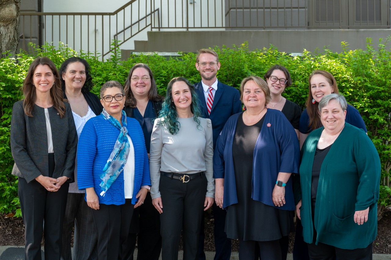 a group shot of the Council staff from spring 2022. It’s ten people posing together outdoors in front of some shrubs, and they are all dressed professionally. There are 9 women and 1 man