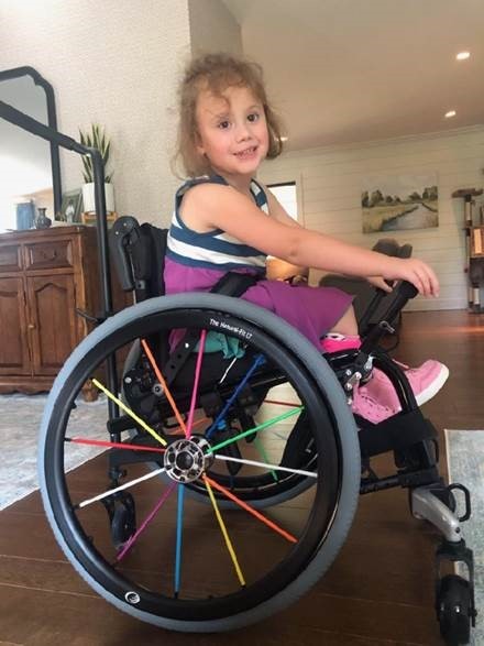 a young little girl with curly hair in her wheelchair in her house, smiling.