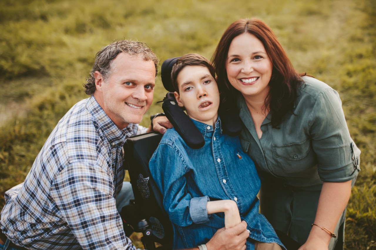 Eli, a young adult with a physical disability seated in his wheelchair with his parents posing with him on either side of him.