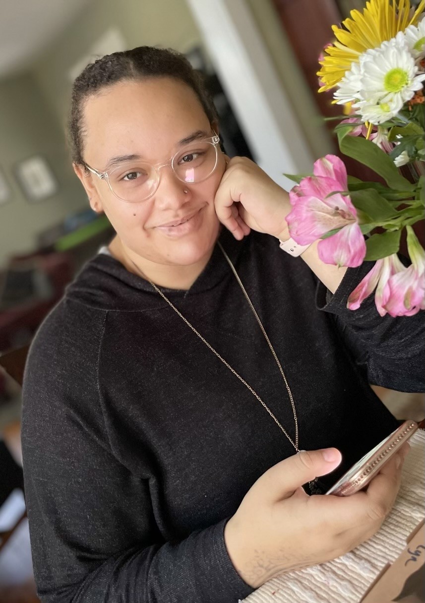 young woman of color sitting at a desk in a black hoodie with her hair pulled back and a nose stud and clear rimmed glasses, with a bouquet of flowers