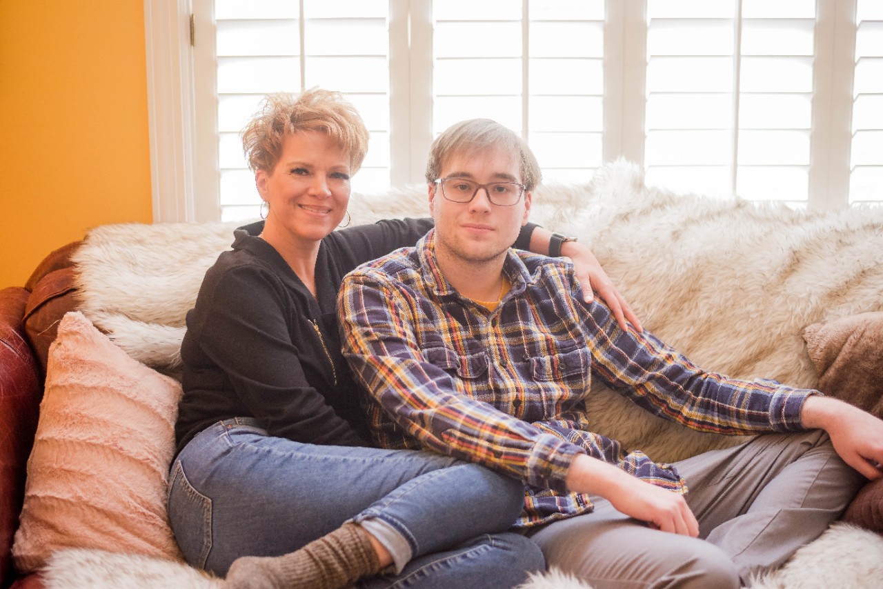 Juli Liske and her son, Ben; white woman with short blonde hair, jeans and a black sweater sits on a couch with her legs curled up next to her and her arm around the shoulders of a young man, her son Ben, who is wearing glasses, a plaid button up shirt, and khakis
