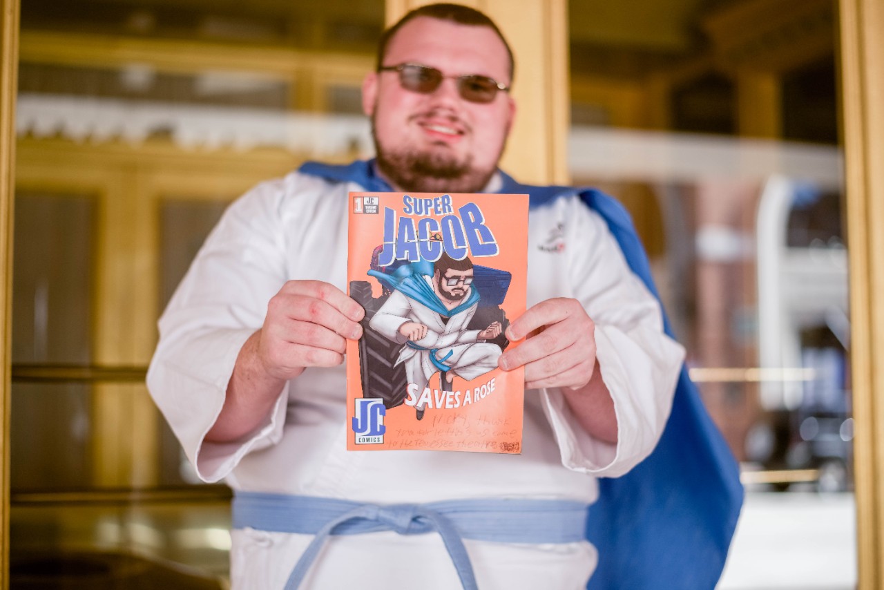 young man with short brown hair and sunglasses and a blue cape, holding up a comic book