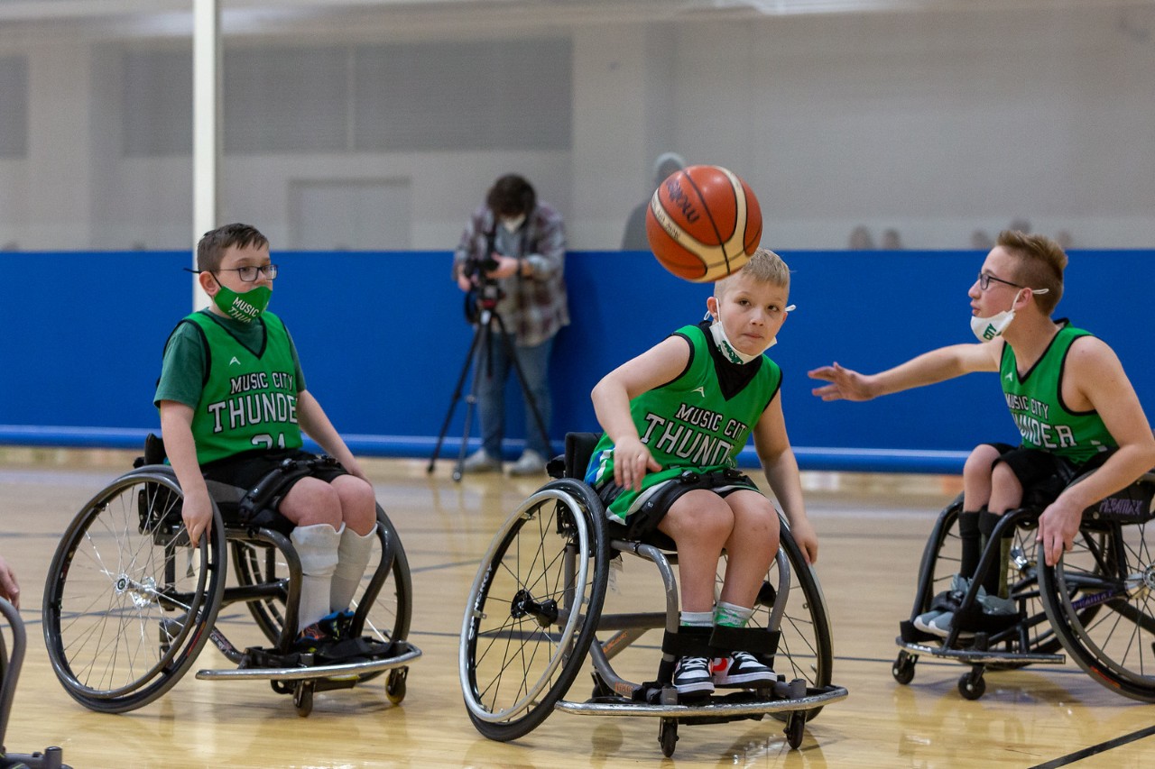3 young boys in green sports jerseys and seated in wheelchairs play basketball in a gym