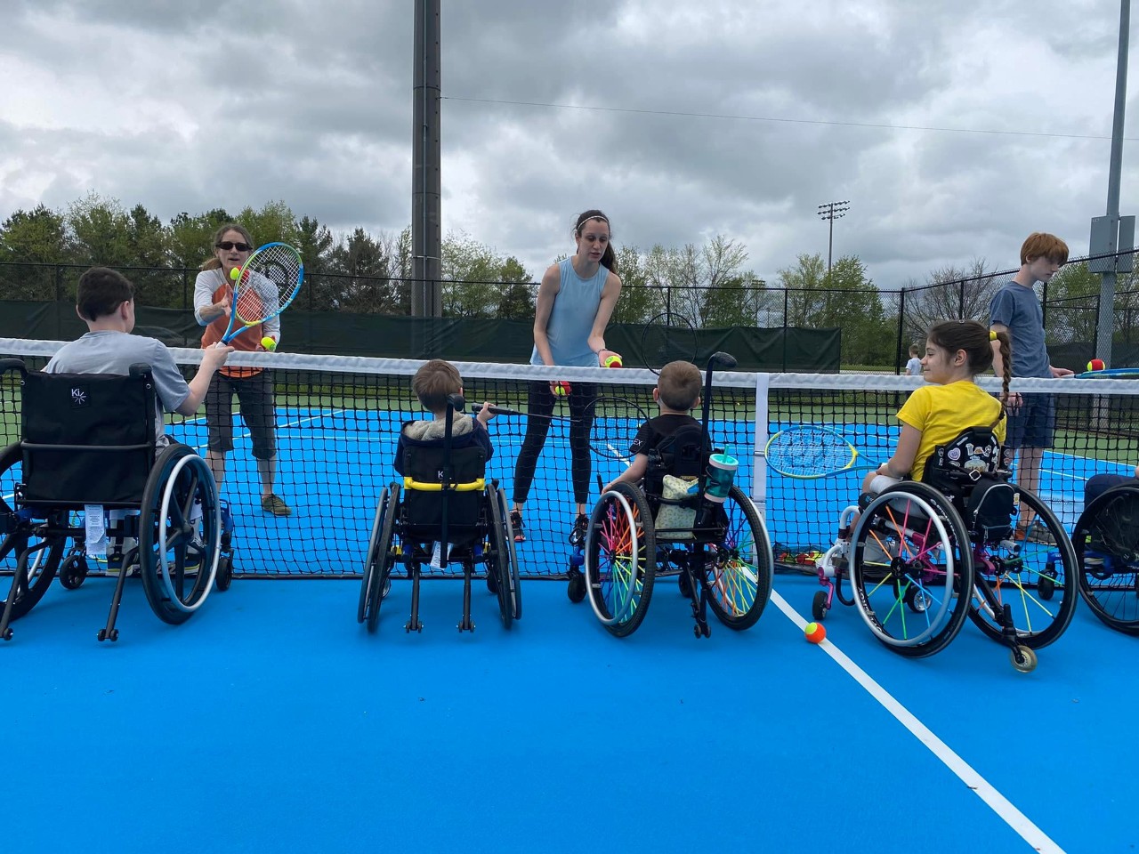 4 kids and young adults are seated in wheelchairs in front of a tennis net on a tennis court holding rackets, facing a couple of instructors