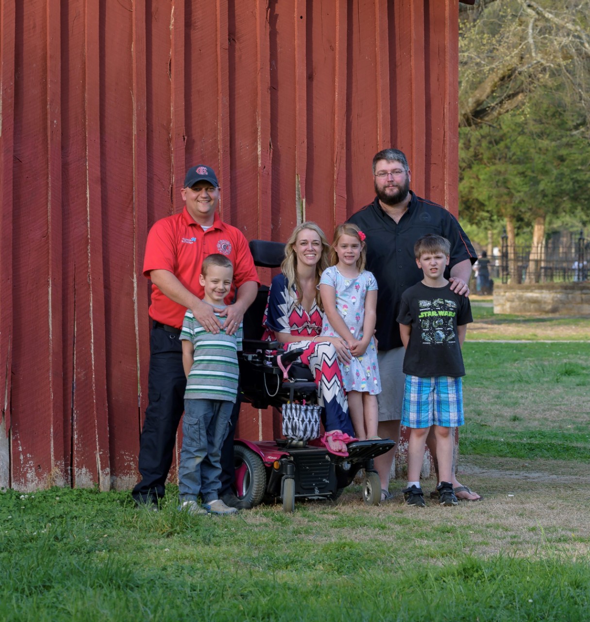 two dads and young sons, and a mom and young daughter pose in front of a red wall at the site of the future inclusive park