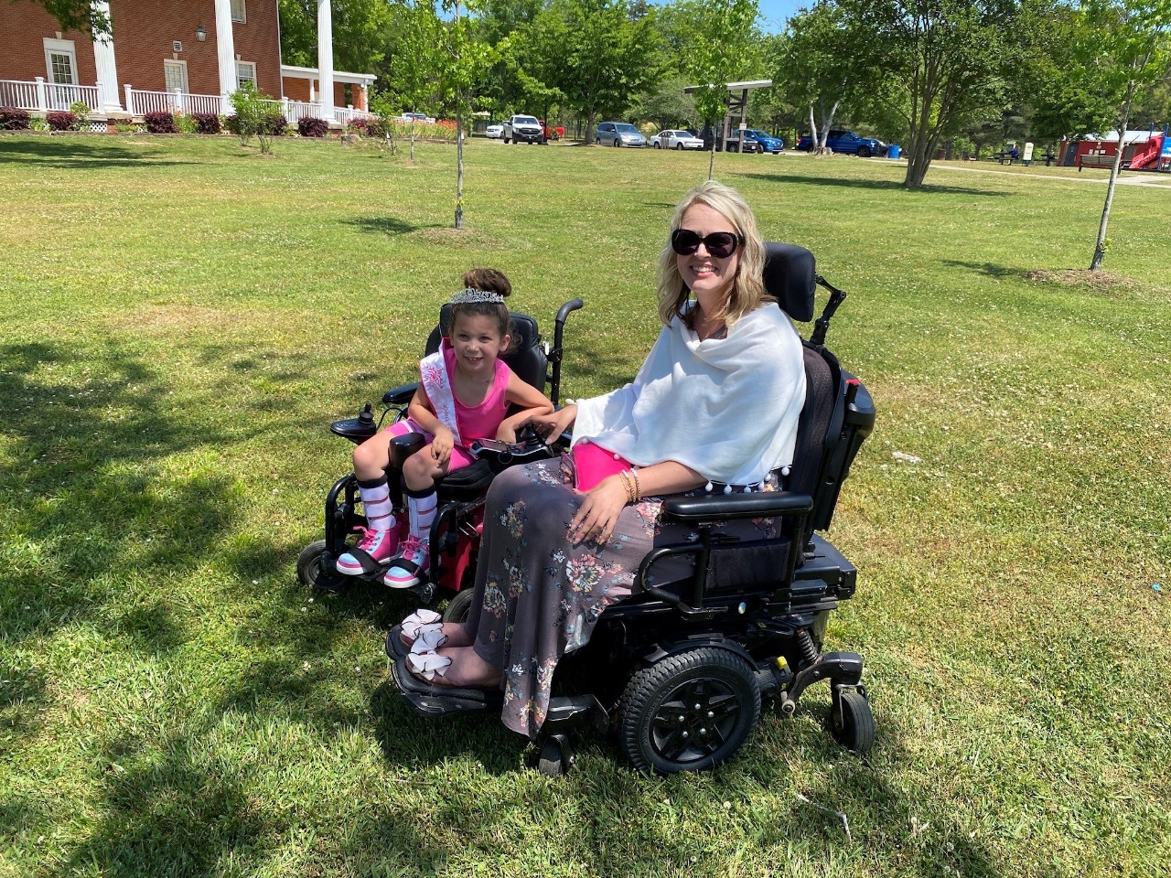 Anora is a young girl of maybe 4-6 years old seated in a wheelchair with a tiara wearing pink and smiling for the photo next to Bliss outdoors; both are in power wheelchairs