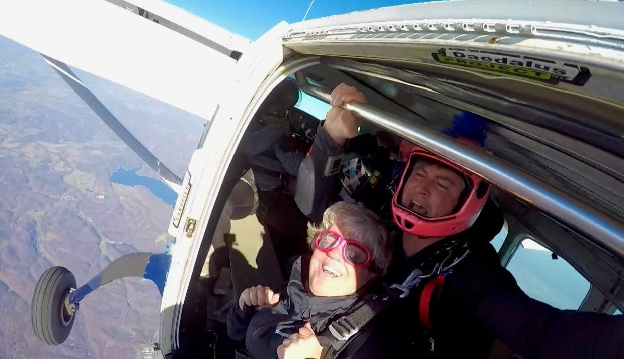 •	Sarah and skydiving instructor at the edge of the plan about to jump out; both are wearing helmets, goggles, gear and harnesses