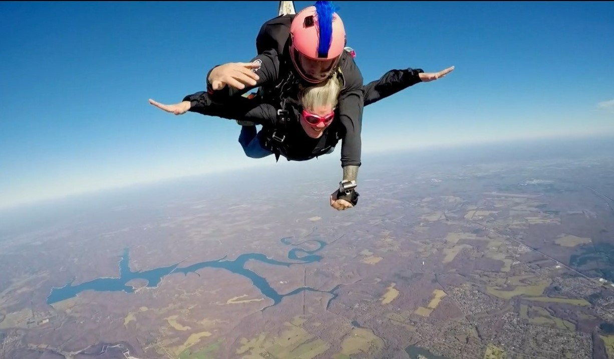 •	A head-on photo of Sarah laying fully parallel to the ground after diving from the plane with her arms outstretched, with the instructor harnessed above her with one arm stretched out below them; an amazing landscape is visible behind and below them