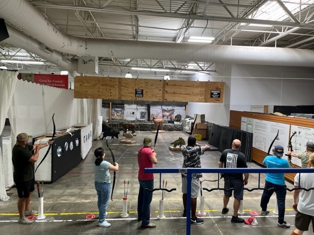 a line of several archers have their backs to the camera in a warehouse space where they are about to shoot arrows at a variety of different animal-shaped targets at varying distances from them