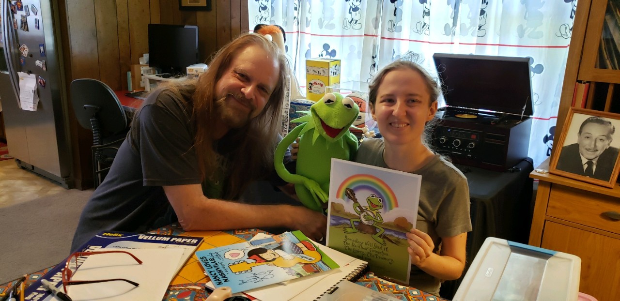 a young white woman with blonde hair is seated next to cartoonist Guy Gilchrist, who is a white man with long blonde hair and a long brown beard; he's holding up a Kermit the Frog puppet between them and Jennifer is holding up a cartoon of Kermit playing the banjo in front of a rainbow