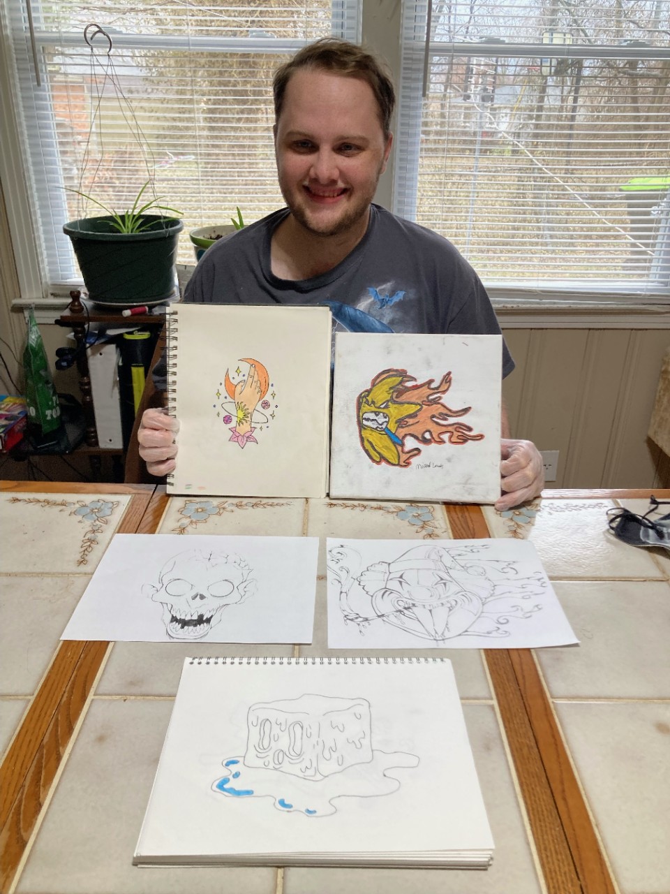 a young white man smiling, holding up a couple of detailed colored sketches and sitting at a table covered in pencil sketches. the sketches on the table include a clown, a melting cube of ice and a skull.