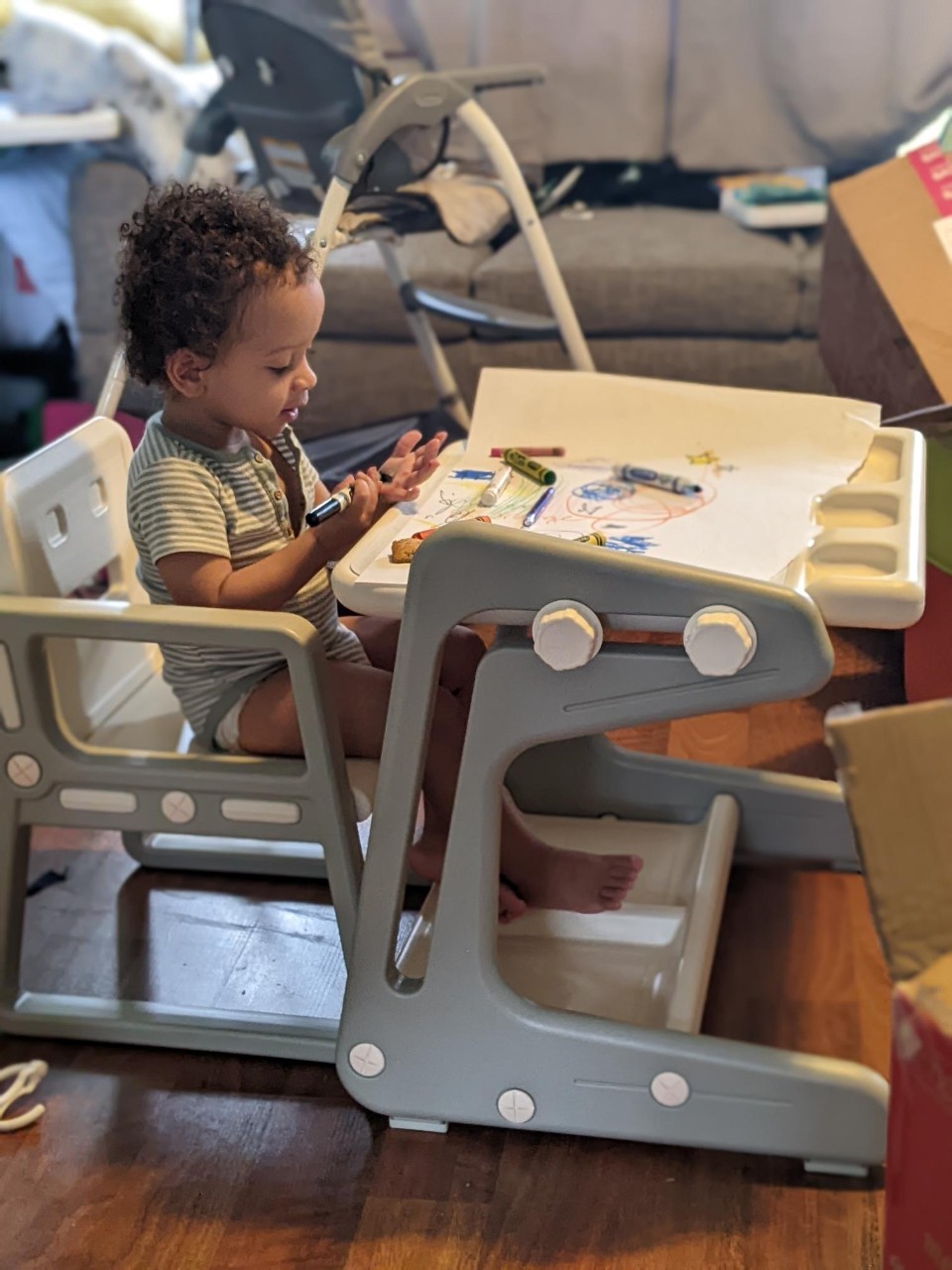 a toddler with curly black hair sits at a child's art table with crayons and paper on the desk in front of him