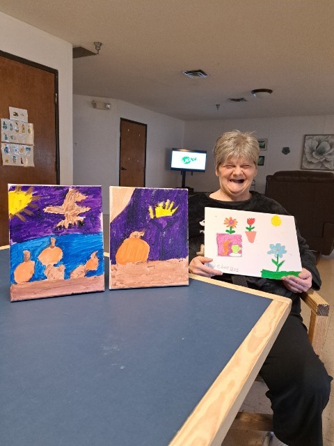 an older white woman with short gray hair and a huge smile sits at a table holding up a painting with two others propped up next to her. the paintings include potted flowers and what appears to be ducks on a body of water and the sun.