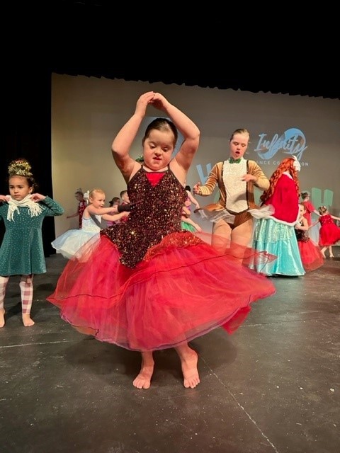 a young girl with down syndrome in a pink dress and large tutu in the middle of a dance move on stage with several other children in costumes